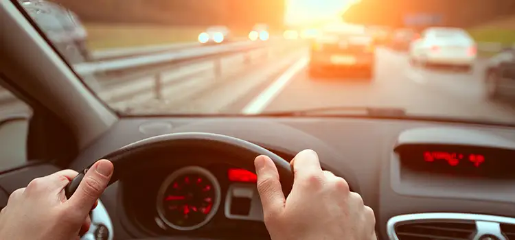 A photo of someone with their hands on the steering wheel while driving towards a sunset on a 4-lane highway.